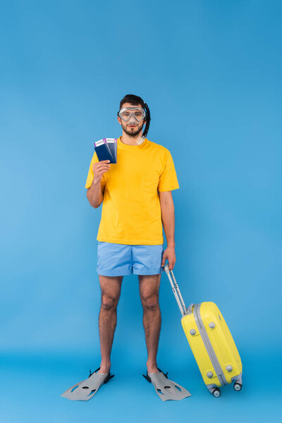 Man in swimming flippers and goggles holding passports and suitcase on blue background 