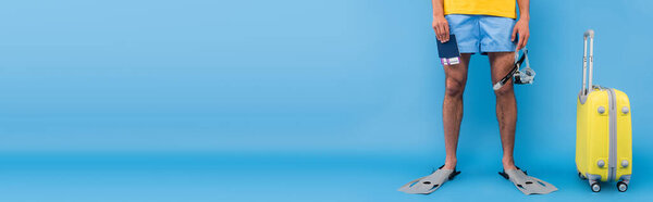 Cropped view of man in swimming flippers holding goggles and passports near suitcase on blue background, banner  