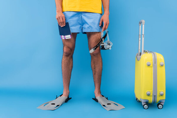 Cropped view of man in swimming flippers holding passports and goggles near suitcase on blue background 