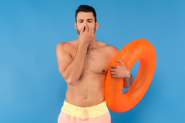 Shirtless man pinching nose and holding swimming ring isolated on blue 