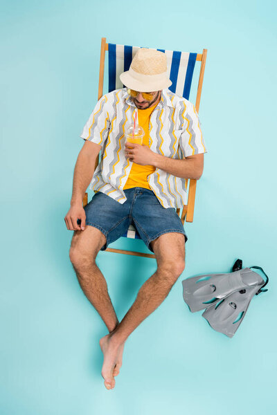 High angle view of man in straw hat holding orange juice near swimming flippers on blue background 