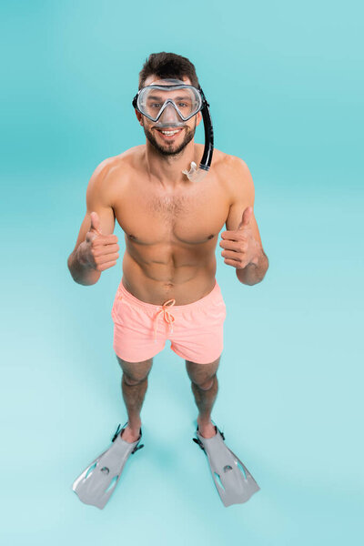 High angle view of shirtless man in swimming goggles and flippers showing like on blue background 