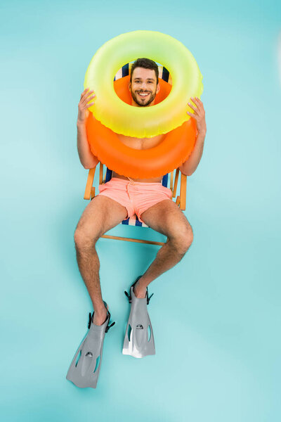 High angle view of smiling man in swimming flippers and inflatable rings sitting on deck chair on blue background 