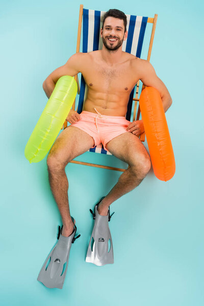High angle view of shirtless man in swimming flippers holding inflatable rings on blue background 