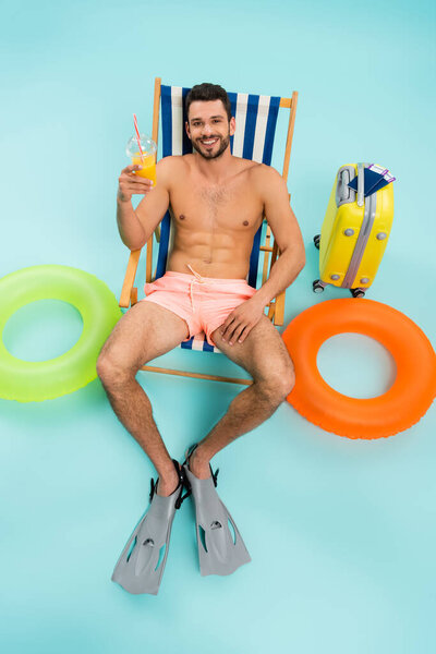High angle view of smiling man in swimming flippers holding orange juice near inflatable rings and suitcase on blue background 