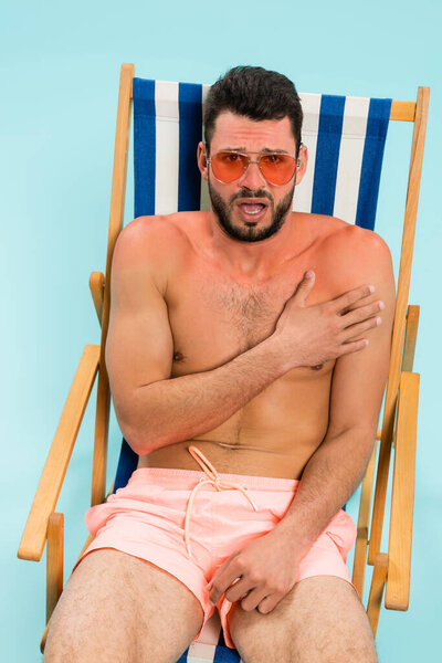 Excited man in sunglasses touching sunburn on deck chair isolated on blue 