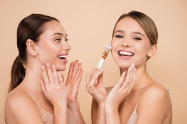 amazed woman looking at laughing friend powdering face isolated on beige clipart