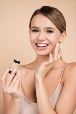 cheerful woman applying face foundation while looking at camera isolated on beige clipart