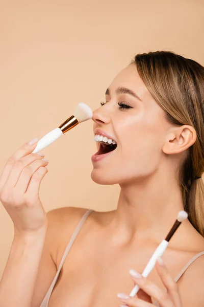 excited woman powdering nose with cosmetic brush isolated on beige