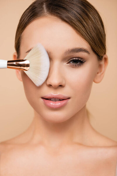 pretty woman with natural visage covering eye with cosmetic brush isolated on beige