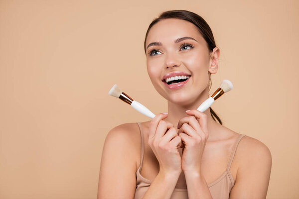 excited woman with cosmetic brushes looking away isolated on beige