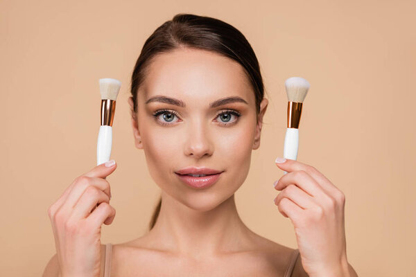 front view of young woman with natural makeup holding cosmetic brushes isolated on beige