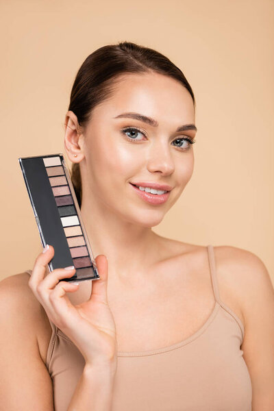pleased woman with eye shadows palette smiling at camera isolated on beige