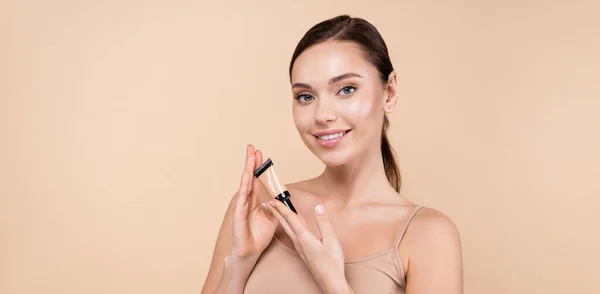 smiling woman with tube of concealer looking at camera isolated on beige, banner