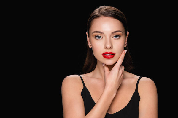 sensual woman with red lips touching face while looking at camera isolated on black
