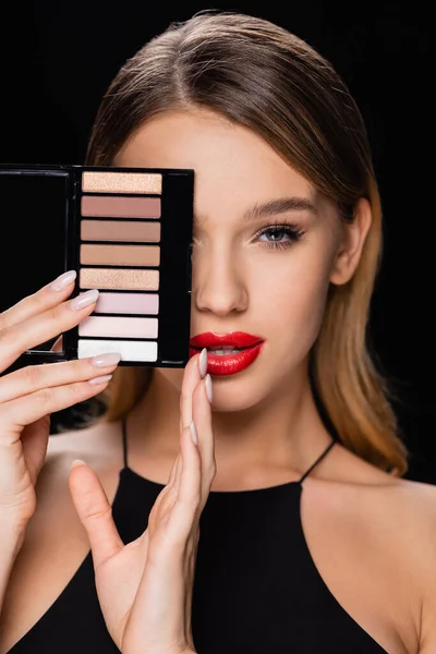 seductive woman obscuring face with eye shadows palette isolated on black