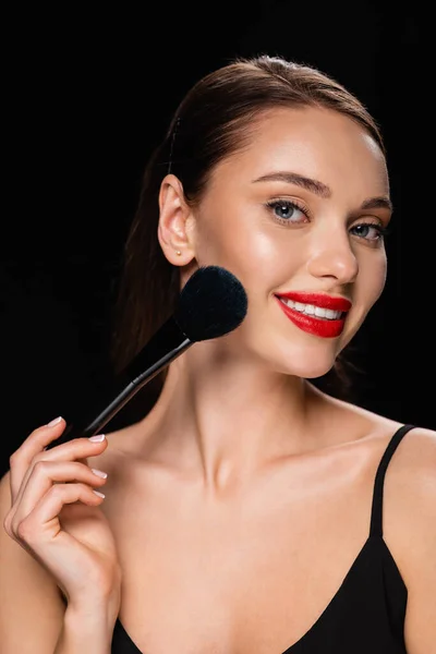 smiling woman powdering face with cosmetic brush isolated on black