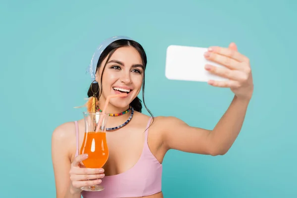 Positive woman with cocktail taking selfie on blurred smartphone isolated on blue