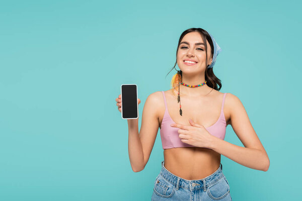 Позитивная женщина сверху указывает на смартфон, изолированный на голубом 