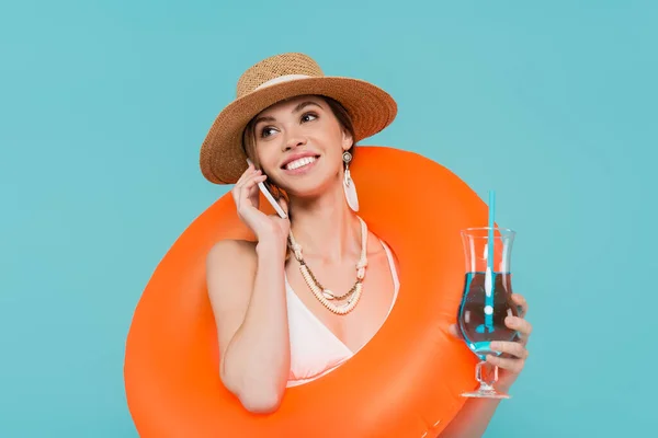 Positive woman with cocktail and swim ring talking on smartphone isolated on blue