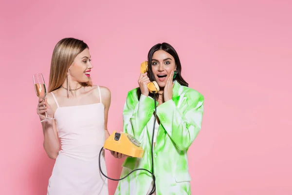 興奮した女性はピンクの背景にシャンパンと友人の近くで電話で話す — ストック写真