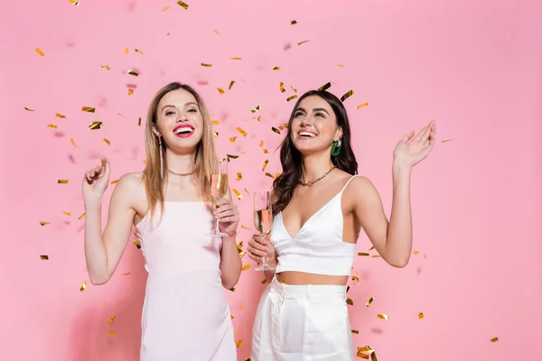 ポジティブなブルネット女性とともにシャンパン立って近く友人とConfetti上のピンクの背景 — ストック写真