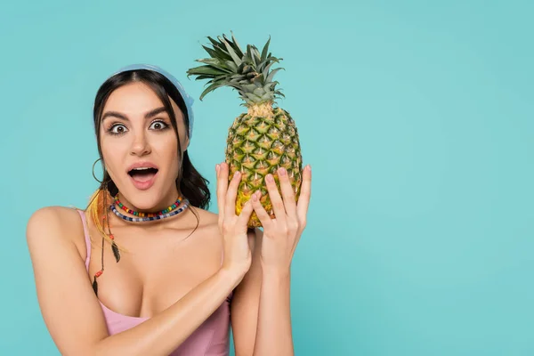 Amazed woman in necklaces holding pineapple isolated on blue