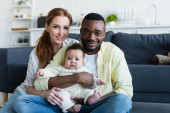 joyful interracial couple with infant child smiling at camera