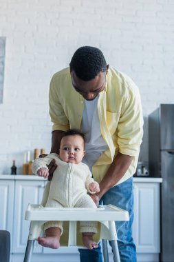 Afrikalı Amerikalı bir adam bebek çocuğunu bebek koltuğunun yanında tutuyor.