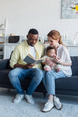 Gülümseyen kadın kanepeye oturmuş bebekle birlikte Afro-Amerikan kocası kitap okuyor.