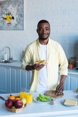 Mutlu Afrikalı Amerikalı adam mutfakta sandviç tutuyor.