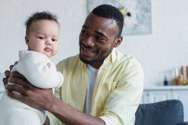 pleased man holding african american baby girl at home clipart