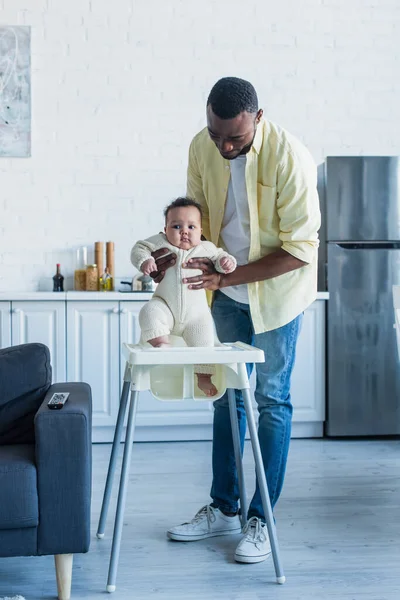 Homem Americano Africano Segurando Criança Infantil Perto Cadeira Bebê Cozinha — Fotografia de Stock