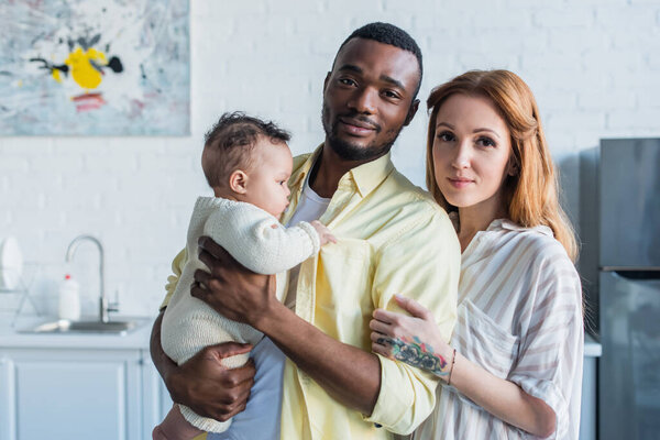african american man holding baby near happy wife at home