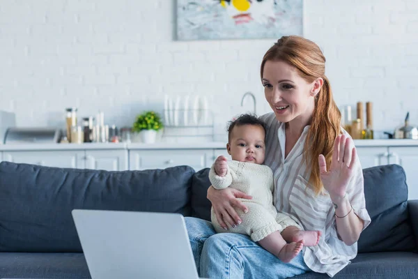 Uśmiechnięta Kobieta Machająca Ręką Przy Laptopie Siedząc Kanapie Afrykańską Córką — Zdjęcie stockowe