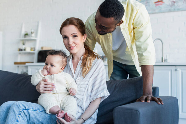 african american man looking at happy wife sitting on sofa with infant daughter