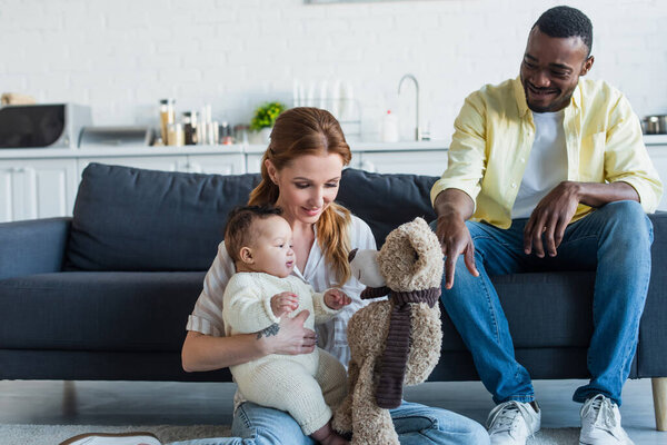 happy woman sitting on floor with infant daughter and teddy bear near smiling african american husband