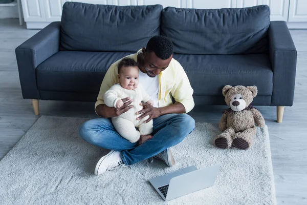 ラップトップとテディベアの近くの床に座っている間に赤ちゃんを抱えているアフリカ系アメリカ人 — ストック写真