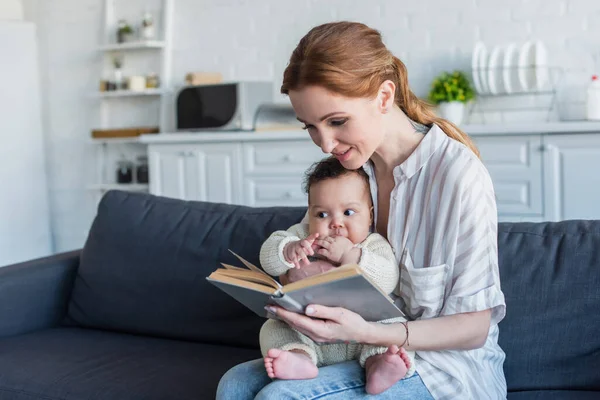 キッチンでソファに座っている間にアフリカ系アメリカ人の女の子に本を読んでいる女性 — ストック写真