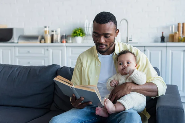 African American Man Reading Book Baby Girl Couch Kitchen — Stock Photo, Image
