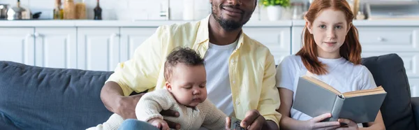 Menina Segurando Livro Perto Pai Americano Africano Irmã Bebê Sofá — Fotografia de Stock