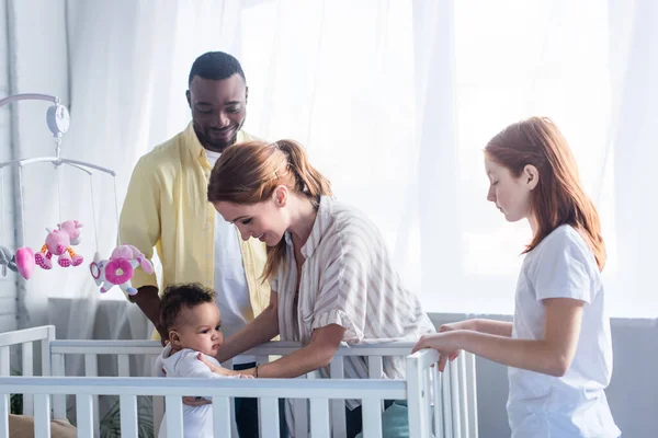 Feliz Mujer Apoyo Bebé Niña Pie Cuna Cerca Familia Multiétnica — Foto de Stock