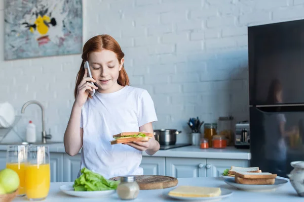 Kızıl Saçlı Kız Sandviçi Tutuyor Mutfakta Cep Telefonuyla Konuşuyor — Stok fotoğraf