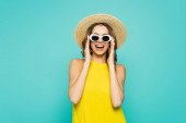 Pozitivní žena v slamáku a sluneční brýle izolované na modré 