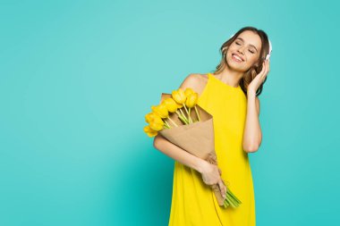 Smiling woman in dress and headphones holding tulips on blue background  clipart
