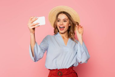 Yaz kıyafetleri ve hasır şapkalı güzel bir kadın pembe arka planda akıllı telefondan selfie çekiyor. 
