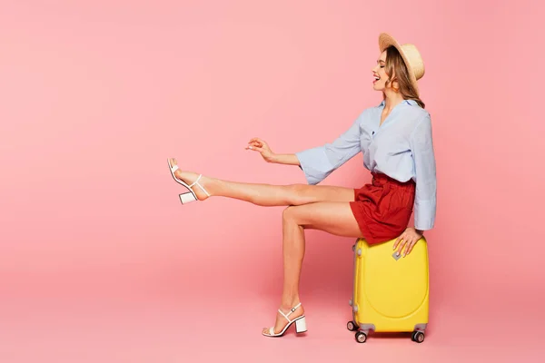 Side view of smiling woman in straw hat sitting on suitcase on pink background
