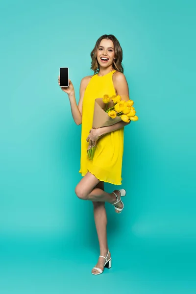 Веселая Женщина Показывает Мобильный Телефон Держит Тюльпаны Синем Фоне — стоковое фото