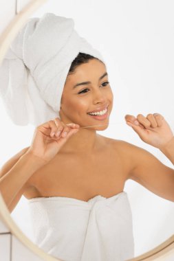 Havluya sarılmış gülümseyen Afro-Amerikan kadın diş ipini yüzüne yakın tutuyor ve banyoda aynaya bakıyor.