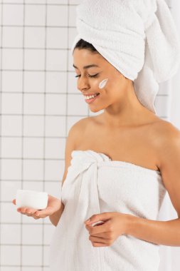 Gülümseyen Afrikalı Amerikalı kadın elinde kremayla kavanoza bakıyor beyaz desenli arka planda